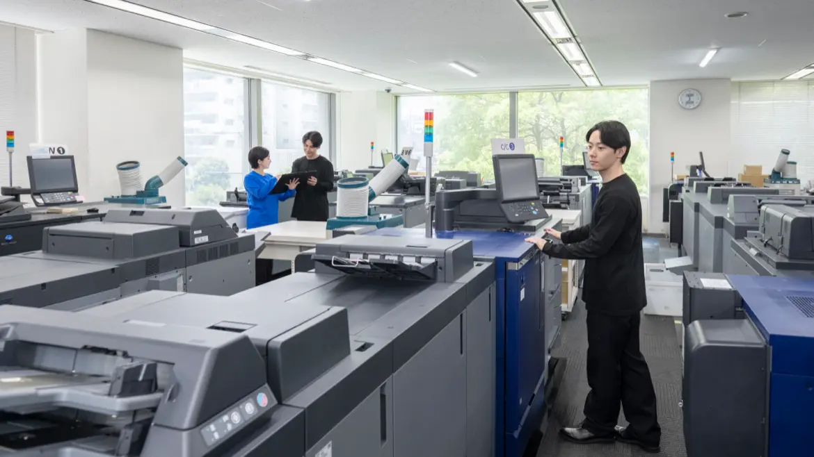 複数のプロダクションプリンターが並ぶ室内で、従業員が印刷作業を行う様子