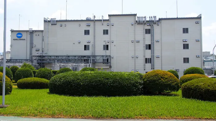 手前の植栽越しに見えるコニカミノルタサプライズ社 甲府本社の白い工場棟の外観写真