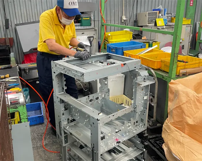協力会社の作業場で、外装を外した複合機の骨組みを作業員が工具で解体している写真