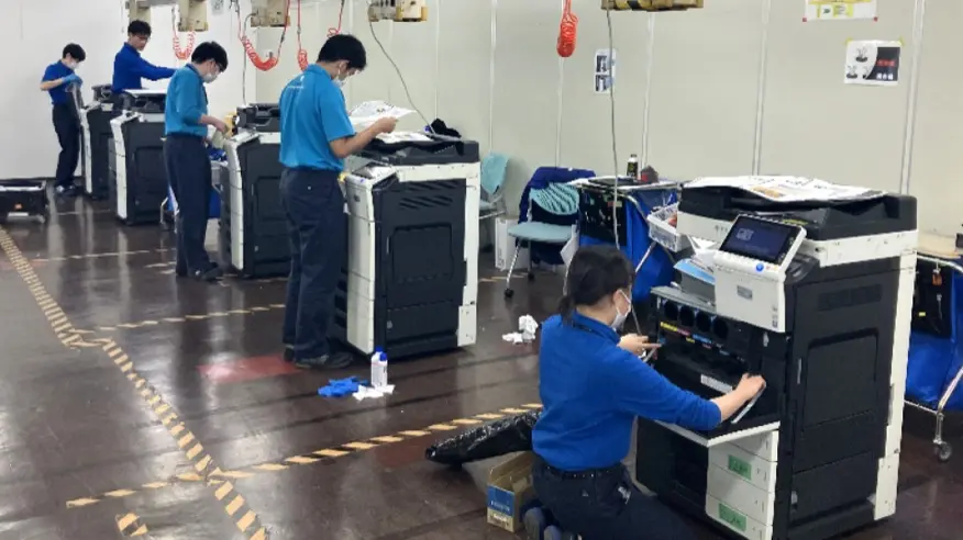 作業場に並べた複数の複合機を、作業員がそれぞれ動作確認・検査している写真