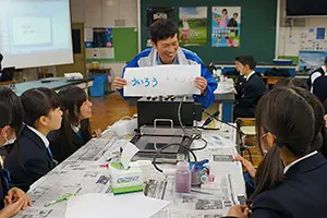 教室で、社員が手動コピーで写し取った紙を生徒に見せている写真