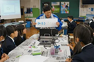 教室で、社員が手動コピーで写し取った紙を生徒に見せている写真